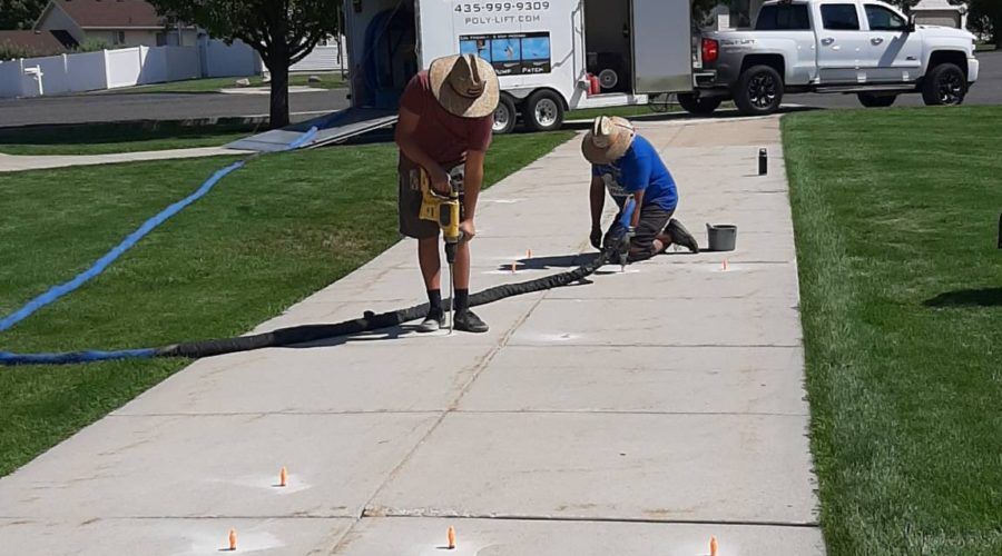 Two Poly-Lift crew members drilling and foam-injecting a settled residential concrete driveway beside the company trailer